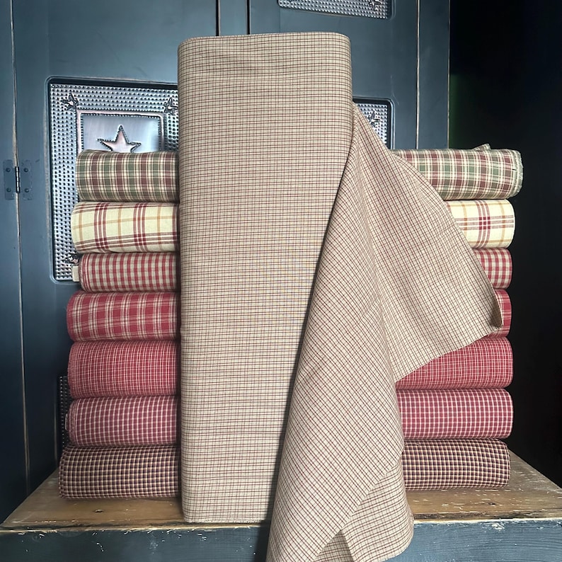 May include: A stack of fabric rolls in various plaid patterns, including red, cream, and brown. A large roll of tan fabric with a grid pattern is in the foreground. The fabrics are stacked on a wooden surface, suggesting a home decor or craft theme.