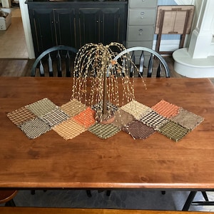 May include: A brown, wooden table with a patchwork table runner in shades of brown, green, and orange. The runner is made of fabric squares with frayed edges. A gold metal decorative tree with lights is in the center of the runner.