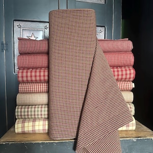 May include: A stack of fabric rolls in various red and tan plaid patterns. A large roll of fabric with a brown and tan check pattern is in the center. The fabrics are displayed on a wooden surface.