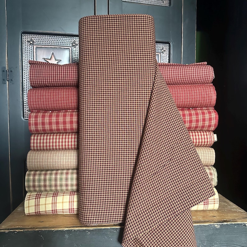 May include: A stack of fabric rolls in various red and tan plaid patterns. A large roll of fabric with a brown and tan check pattern is in the center. The fabrics are displayed on a wooden surface.