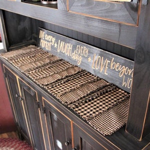 May include: A black cabinet with a wooden sign that reads "Live every moment, Laugh every day, Love beyond words." The cabinet has a series of black and tan checkered placemats on top.
