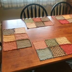 K&ouml;nnte beinhalten: Vier Patchwork-Platzsets mit ausgefransten Kanten in Rot-, Gr&uuml;n-, Blau- und Braunt&ouml;nen auf einem Holztisch.