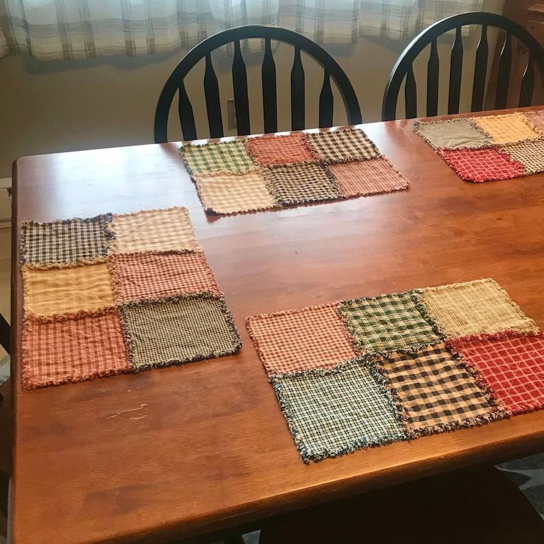 K&ouml;nnte beinhalten: Vier Patchwork-Platzsets mit ausgefransten Kanten in Rot-, Gr&uuml;n-, Blau- und Braunt&ouml;nen auf einem Holztisch.