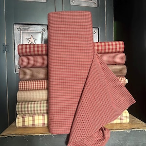 May include: A stack of fabric bolts in various plaid patterns, with a large roll of red and white checked fabric in the foreground. The fabrics are in shades of red, tan, and green, suggesting a country or farmhouse style.