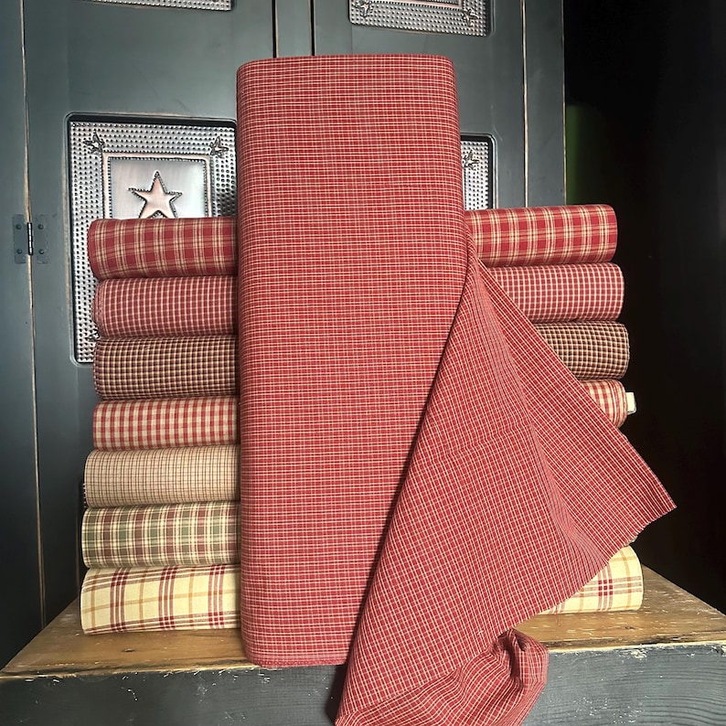 May include: A stack of fabric bolts in various plaid patterns, with a large roll of red and white checked fabric in the foreground. The fabrics are in shades of red, tan, and green, suggesting a country or farmhouse style.