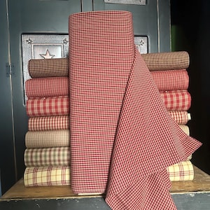 May include: A stack of various fabric rolls in shades of red, brown, and cream. The fabrics feature plaid and check patterns. A large roll of red and white checked fabric is in the foreground, with a portion draped over the stack. The fabrics are displayed on a wooden surface.