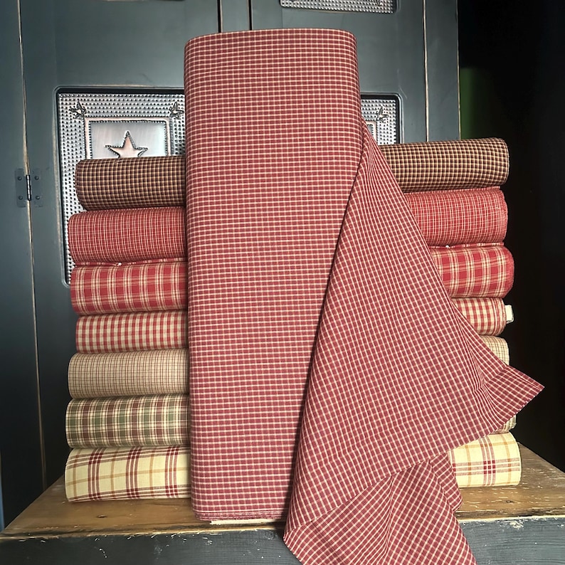 May include: A stack of various fabric rolls in shades of red, brown, and cream. The fabrics feature plaid and check patterns. A large roll of red and white checked fabric is in the foreground, with a portion draped over the stack. The fabrics are displayed on a wooden surface.