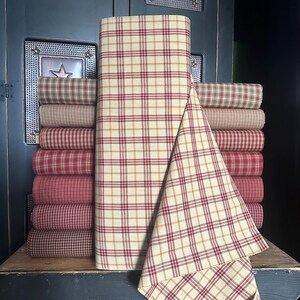 May include: A stack of rolled fabric bolts in various plaid patterns. The fabrics feature a cream background with red, brown, and gold plaid designs. The fabrics are stacked on a wooden surface.