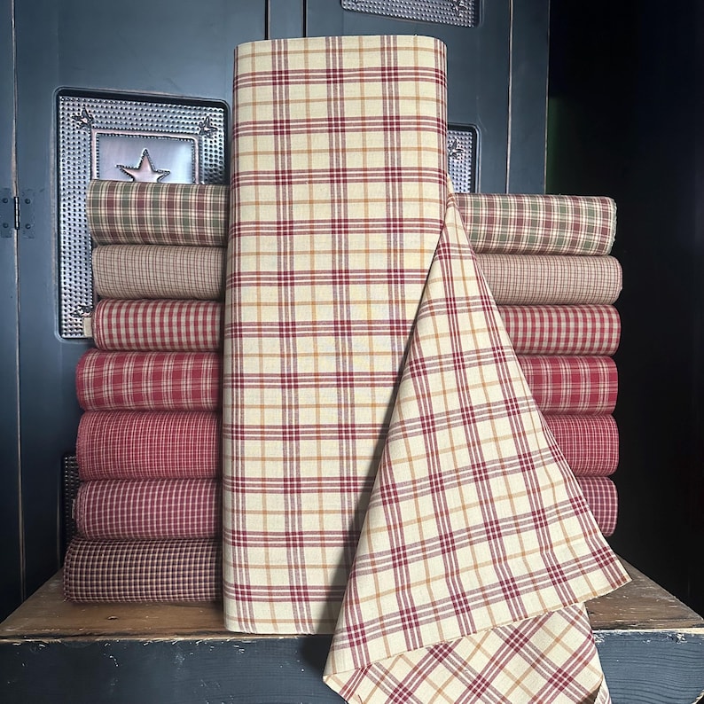 May include: A stack of rolled fabric bolts in various plaid patterns. The fabrics feature a cream background with red, brown, and gold plaid designs. The fabrics are stacked on a wooden surface.