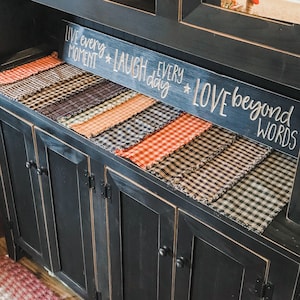 May include: Black painted cabinet with multiple drawers and doors. The cabinet is decorated with a variety of plaid fabric runners with frayed edges. A sign above the runners reads "Live every moment * laugh every day * love beyond words".