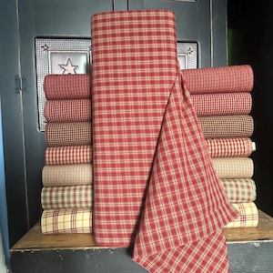 May include: A stack of fabric rolls in various plaid patterns and colors, including red, tan, and brown. A large roll of red and tan plaid fabric is in the center. The fabrics are arranged on a wooden surface.