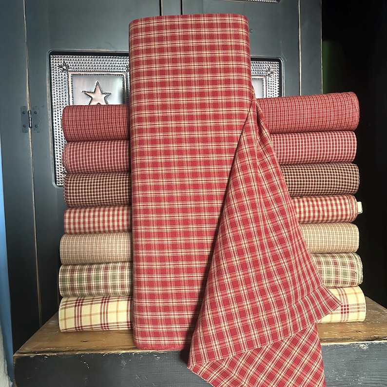 May include: A stack of fabric rolls in various plaid patterns and colors, including red, tan, and brown. A large roll of red and tan plaid fabric is in the center. The fabrics are arranged on a wooden surface.