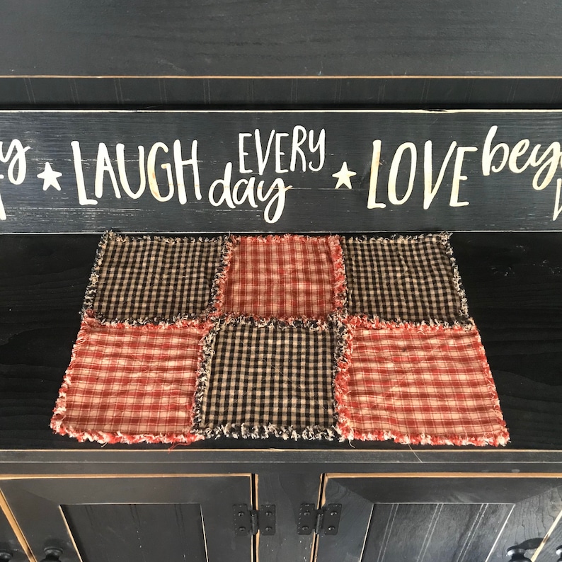 K&ouml;nnte beinhalten: Ein roter und schwarzer karierter Tischl&auml;ufer mit ausgefransten Kanten. Der Tischl&auml;ufer ist aus Stoff und hat ein Patchwork-Design. Der Tischl&auml;ufer liegt auf einer schwarzen Oberfl&auml;che. Der Text "Laugh every day Love begins" ist auf einem Schild &uuml;ber dem Tischl&auml;ufer zu sehen.