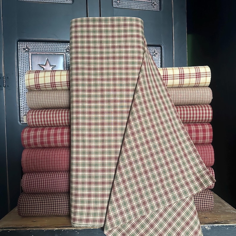 May include: A stack of fabric rolls in various plaid patterns. The dominant roll features a green, red, and cream plaid, with other rolls in shades of red, cream, and brown. The fabric is likely cotton or a similar material, suitable for quilting or crafting projects.