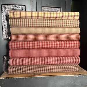 May include: A stack of rolled fabric bolts in various plaid patterns. The colors include red, tan, and green, with a mix of light and dark shades. The fabric is stacked on a wooden surface, suggesting a craft or textile theme.
