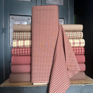 May include: A stack of various fabric rolls in a variety of colors and patterns. The fabrics include red and white gingham, plaid, and solid colors. The fabrics are stacked on a wooden surface against a dark blue background.
