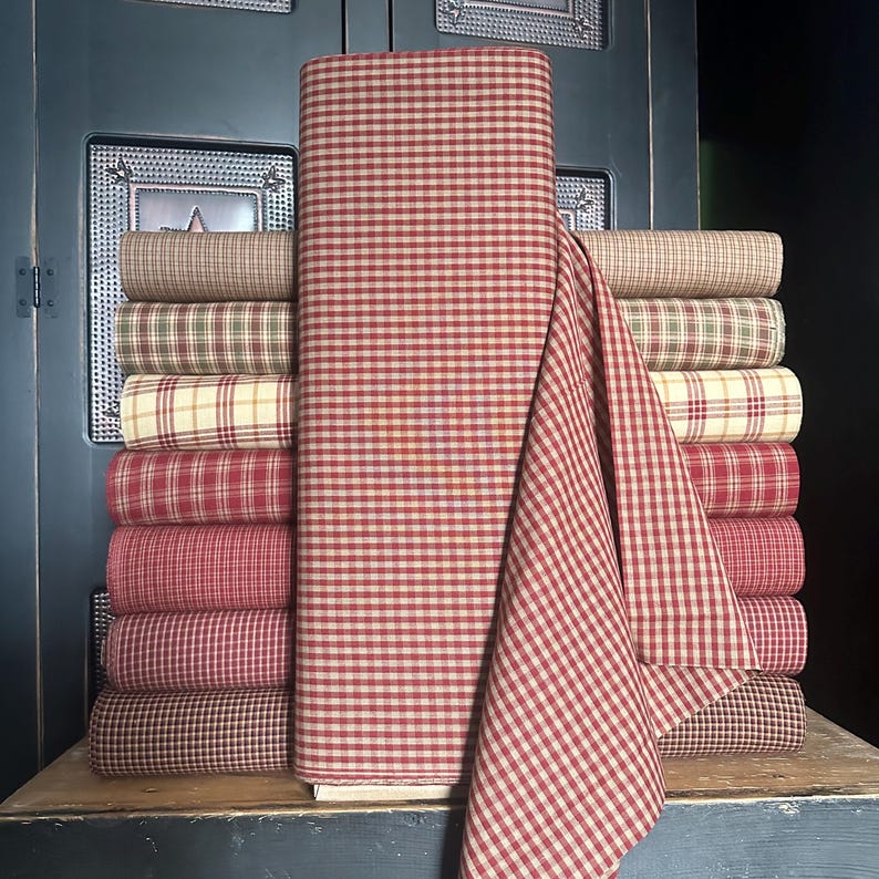 May include: A stack of various fabric rolls in a variety of colors and patterns. The fabrics include red and white gingham, plaid, and solid colors. The fabrics are stacked on a wooden surface against a dark blue background.