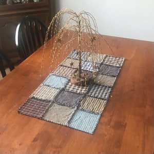 May include: A brown wooden table with a patchwork quilt table runner featuring a variety of blue, brown, and beige squares. A gold metal willow tree sculpture is centered on the table runner.