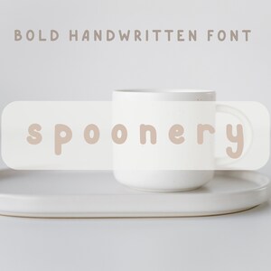 May include: A white ceramic mug with a speckled finish, sitting on a white oval tray. The word "spoonery" is written in a bold, handwritten font on a white rectangle behind the mug.