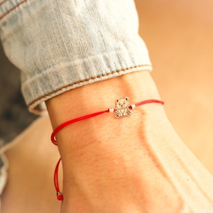 May include: Red string bracelet featuring a small silver-colored teddy bear charm. The charm has a heart-shaped detail. The bracelet is worn on a wrist, with a denim jacket sleeve visible in the background.