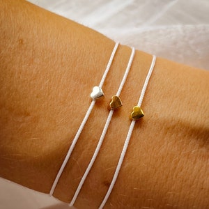 Peut inclure: Trois bracelets en cordon blanc avec des breloques en forme de cœur. Les breloques sont en argent, en or et en or.