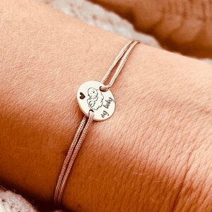 Puede incluir: Un brazalete de cordón beige con un dije de plata que dice "my baby" y tiene un pequeño corazón y un dibujo de un bebé en una nube.