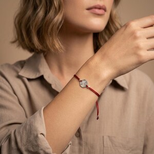 Könnte beinhalten: Ein rotes Kordelarmband mit einem silberfarbenen, kreisförmigen Anhänger mit Weltkarten-Design. Das Armband wird am Handgelenk getragen, der Anhänger mittig auf der roten Kordel.