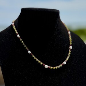May include: A necklace featuring a thin, braided cord strung with small, round, olive-green beads, interspersed with tiny, dark-red accents and irregular, white, lustrous pearls. The beads and pearls are arranged in a repeating pattern along the cord.