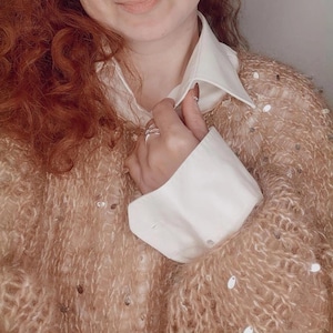 May include: A tan, open-weave sweater with scattered sequins is worn over a white collared shirt. The sweater's texture is visible, and the white cuffs of the shirt are buttoned. The image is a close-up of the clothing.