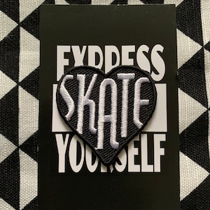 May include: A black heart-shaped patch embroidered with the word "SKATE" in white, on a black card with the text "EXPRESS YOURSELF" in white. The background features a black and white geometric pattern.