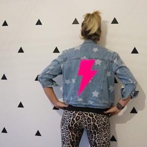 May include: A light-wash denim jacket with white star patterns and a large, bright pink lightning bolt graphic on the back. The jacket has rolled-up sleeves. The person is wearing leopard print leggings.
