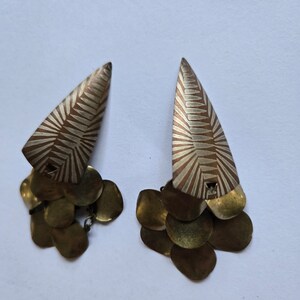 Puede incluir: Un par de pendientes de oro con un diseño geométrico. Los pendientes están hechos de dos partes: una parte superior triangular y un grupo de colgantes redondos.