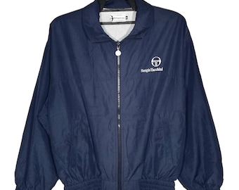 May include: A navy blue windbreaker jacket featuring a full-length front zipper, a fold-down collar, and long sleeves with elastic cuffs. The Sergio Tacchini logo and text are embroidered in white on the upper left chest area of the garment.