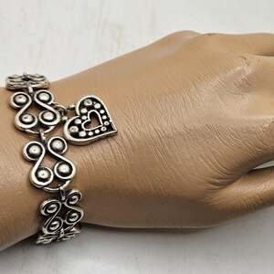 Puede incluir: Pulsera plateada con un dije de coraz&oacute;n y adornos decorativos. La pulsera se muestra en una mano de maniqu&iacute;. El dije de coraz&oacute;n tiene un dise&ntilde;o recortado y est&aacute; acentuado con peque&ntilde;os puntos. La pulsera tiene un patr&oacute;n repetitivo.