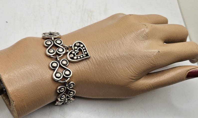 Puede incluir: Pulsera plateada con un dije de coraz&oacute;n y adornos decorativos. La pulsera se muestra en una mano de maniqu&iacute;. El dije de coraz&oacute;n tiene un dise&ntilde;o recortado y est&aacute; acentuado con peque&ntilde;os puntos. La pulsera tiene un patr&oacute;n repetitivo.