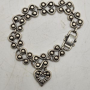 Puede incluir: Pulsera plateada con un patr&oacute;n repetitivo de espirales conectadas y peque&ntilde;as esferas. Un dije en forma de coraz&oacute;n con un recorte de coraz&oacute;n y borde punteado cuelga de la pulsera. La pulsera tiene un cierre.
