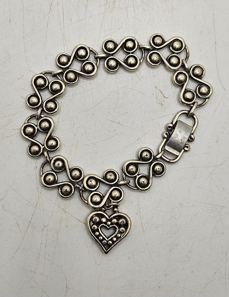 Puede incluir: Pulsera plateada con un patr&oacute;n repetitivo de espirales conectadas y peque&ntilde;as esferas. Un dije en forma de coraz&oacute;n con un recorte de coraz&oacute;n y borde punteado cuelga de la pulsera. La pulsera tiene un cierre.