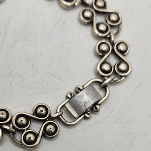 Puede incluir: Pulsera de metal plateado con un patr&oacute;n repetitivo de remolinos estilizados y acentos esf&eacute;ricos. La pulsera tiene un cierre rectangular y probablemente sea vintage o est&eacute; inspirada en dise&ntilde;os retro. El dise&ntilde;o es sim&eacute;trico y detallado.
