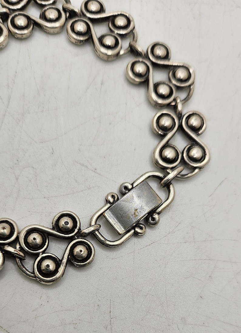 Puede incluir: Pulsera de metal plateado con un patr&oacute;n repetitivo de remolinos estilizados y acentos esf&eacute;ricos. La pulsera tiene un cierre rectangular y probablemente sea vintage o est&eacute; inspirada en dise&ntilde;os retro. El dise&ntilde;o es sim&eacute;trico y detallado.