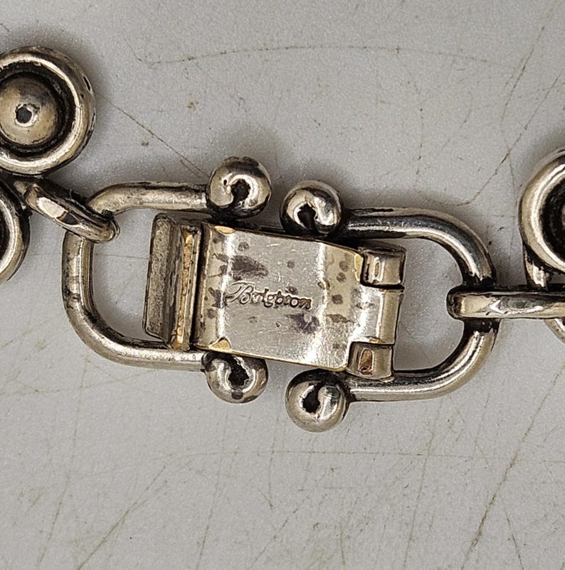 Puede incluir: Primer plano de un cierre de joyer&iacute;a plateado con el nombre de la marca "Brighton" grabado. El cierre est&aacute; conectado a una cadena con eslabones circulares decorativos.