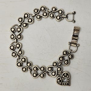Puede incluir: Pulsera plateada con un patr&oacute;n repetitivo de espirales y c&iacute;rculos conectados. Un dije en forma de coraz&oacute;n con un borde punteado y un recorte de coraz&oacute;n cuelga de la pulsera. Cierre de broche.