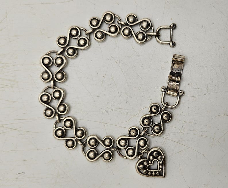 Puede incluir: Pulsera plateada con un patr&oacute;n repetitivo de espirales y c&iacute;rculos conectados. Un dije en forma de coraz&oacute;n con un borde punteado y un recorte de coraz&oacute;n cuelga de la pulsera. Cierre de broche.