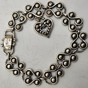Puede incluir: Pulsera plateada con un patr&oacute;n repetitivo de espirales y esferas estilizadas. Un dije en forma de coraz&oacute;n con un centro recortado cuelga de la pulsera. La pulsera tiene un cierre.
