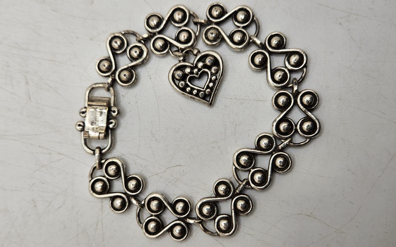 Puede incluir: Pulsera plateada con un patr&oacute;n repetitivo de espirales y esferas estilizadas. Un dije en forma de coraz&oacute;n con un centro recortado cuelga de la pulsera. La pulsera tiene un cierre.