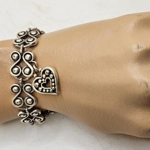 Puede incluir: Pulsera plateada con un dise&ntilde;o ornamentado y un dije en forma de coraz&oacute;n. La pulsera est&aacute; compuesta por elementos decorativos repetidos y un colgante de coraz&oacute;n con un borde punteado y un recorte de coraz&oacute;n. La joya se exhibe en una mano de maniqu&iacute;.