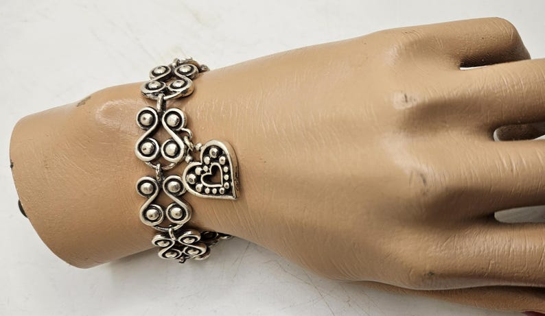 Puede incluir: Pulsera plateada con un dise&ntilde;o ornamentado y un dije en forma de coraz&oacute;n. La pulsera est&aacute; compuesta por elementos decorativos repetidos y un colgante de coraz&oacute;n con un borde punteado y un recorte de coraz&oacute;n. La joya se exhibe en una mano de maniqu&iacute;.
