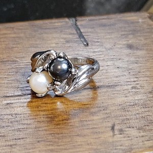 May include: A silver-toned metal ring featuring two round pearls, one white and one dark gray, set within a polished, swirling metal band. The design showcases a bypass style where the two pearls are positioned side-by-side in a decorative setting.