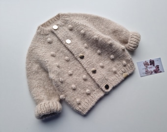 May include: A beige knitted cardigan with a textured bobble stitch and gold buttons. The cardigan has long sleeves and a ribbed trim.