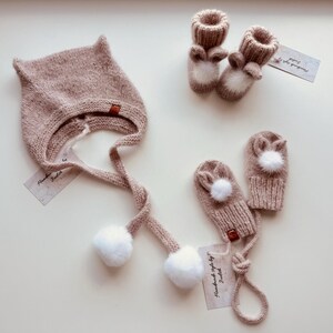 May include: A matching set of knitted baby accessories in a soft pink hue. The set includes a hat with ear-like points, a pair of mittens, and a pair of booties, all adorned with white pom-poms and bunny ear details. Each item has a small tag.