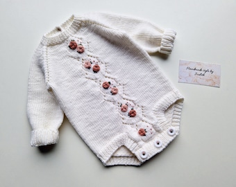 May include: Cream-coloured knitted baby romper with long sleeves and floral detailing. The romper has a diamond-shaped pattern with embroidered pink roses and small white buttons. The tag reads "Handmade style by Judith."